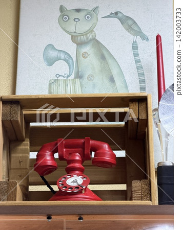 Kolomna, Russia - April 06, 2026: Vintage red rotary telephone sits on a wooden crate with a cat illustration and decorative items visible in the background Kolomna, Russia - April 06, 2026: Vintage red rotary telephone sits on a wooden crate with a cat illustration and decorative items visible in the background 142003733