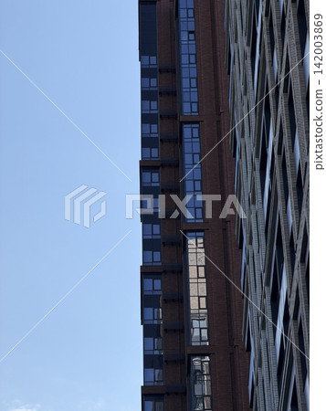 Modern high-rise residential building features glass windows and a brick facade, set against a clear blue sky in an urban environment Modern high-rise residential building features glass windows and a brick facade, set against a clear blue sky in an urban environment 142003869