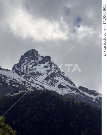 Snow-covered mountain peak towers over lush green forest, with dramatic clouds creating a striking backdrop in an alpine environment Snow-covered mountain peak towers over lush green forest, with dramatic clouds creating a striking backdrop in an alpine environment 142003906