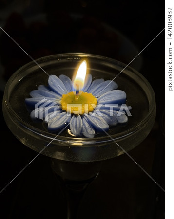 Blue flower-shaped candle with yellow center is lit in a glass holder, casting a warm glow against a dark background with blurred objects 142003932