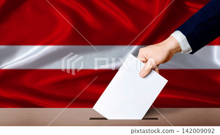 Hand placing a ballot in ballot box with the Latvian flag in the background. Latvia Parliamentary vote 2026 Hand placing a ballot in ballot box with the Latvian flag in the background. Latvia Parliamentary vote 2026 142009092