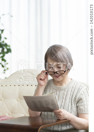 Senior woman reading a newspaper wearing reading glasses in the living room Senior woman reading a newspaper wearing reading glasses in the living room 142010173