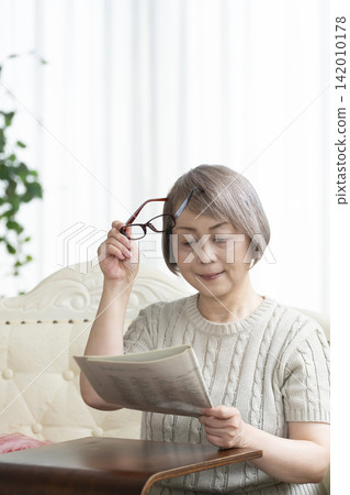 一位老婦人在客廳裡戴著老花眼鏡看報紙。 一位老婦人在客廳裡戴著老花眼鏡看報紙。 142010178