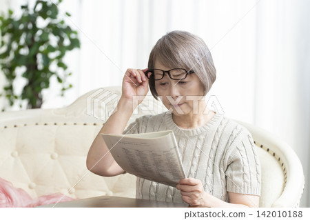 Senior woman reading a newspaper wearing reading glasses in the living room Senior woman reading a newspaper wearing reading glasses in the living room 142010188