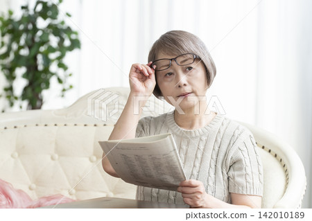 Senior woman reading a newspaper wearing reading glasses in the living room Senior woman reading a newspaper wearing reading glasses in the living room 142010189