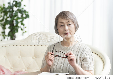 Senior woman reading a newspaper wearing reading glasses in the living room Senior woman reading a newspaper wearing reading glasses in the living room 142010195
