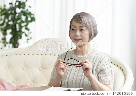 Senior woman reading a newspaper wearing reading glasses in the living room 142010196