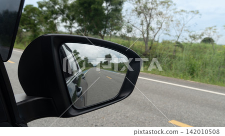 Car side mirror reflecting an asphalt road. Stretching into the distance under an open sky. Beautiful green trees and grass along the roadside. Car side mirror reflecting an asphalt road. Stretching into the distance under an open sky. Beautiful green trees and grass along the roadside. 142010508