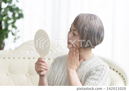 A senior woman holding a hand mirror in her living room 142011020