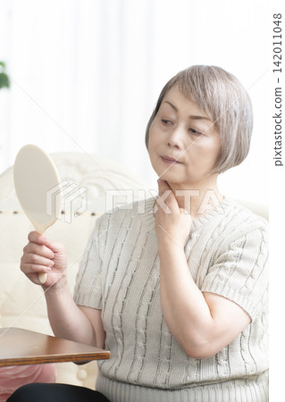 A senior woman holding a hand mirror in her living room A senior woman holding a hand mirror in her living room 142011048