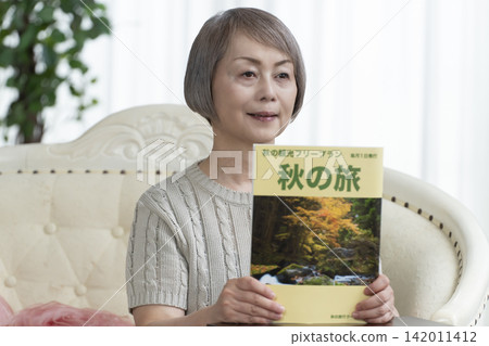 A senior woman is looking at a travel magazine on the living room sofa. A senior woman is looking at a travel magazine on the living room sofa. 142011412