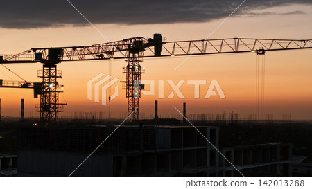 Construction cranes silhouetted against a vibrant sunset sky Construction cranes silhouetted against a vibrant sunset sky 142013288