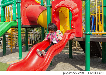 Cheerful little girl sliding down a red slide at outdoor colorful playground Cheerful little girl sliding down a red slide at outdoor colorful playground 142014370