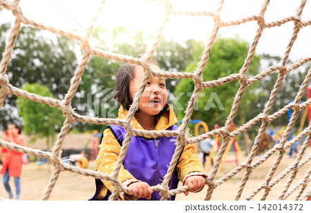 Excited Asian little girl climbing a rope net at an outdoor playground, focus and physical activity, kids outdoor adventure and development. 142014372