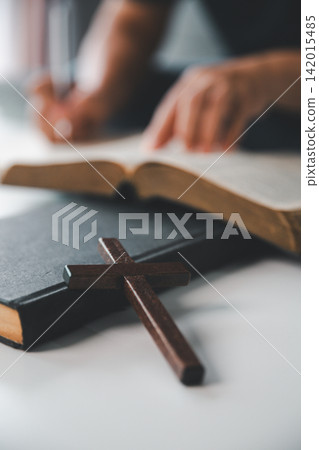 Wooden Christian cross beside an open Holy Bible while a person writes study notes in the background, representing scripture study, devotion, learning, faith, and spiritual growth. Wooden Christian cross beside an open Holy Bible while a person writes study notes in the background, representing scripture study, devotion, learning, faith, and spiritual growth. 142015485