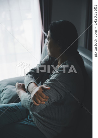 Lonely adult woman hugging herself while sitting beside a window in a dark room, expressing sadness, anxiety, emotional isolation, insecurity, and a need for comfort. 142015603