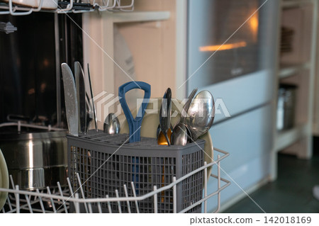 Cutlery basket filled with clean spoons, knives and kitchen utensils inside open dishwasher. Housework, dishwashing, kitchen cleaning, household routine, organization, hygiene, domestic life Cutlery basket filled with clean spoons, knives and kitchen utensils inside open dishwasher. Housework, dishwashing, kitchen cleaning, household routine, organization, hygiene, domestic life 142018169