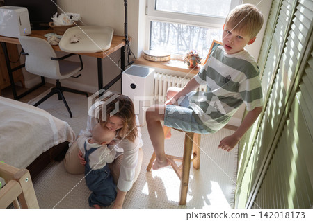 Mother cuddles baby son on floor while older brother sits nearby during relaxed family moment at home. Motherhood, sibling bond, childcare, parenting, affection, family care, childhood, togetherness Mother cuddles baby son on floor while older brother sits nearby during relaxed family moment at home. Motherhood, sibling bond, childcare, parenting, affection, family care, childhood, togetherness 142018173