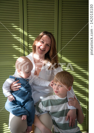 Smiling mother embraces two sons while baby brother reaches toward older sibling during warm family moment at home. Motherhood, sibling bond, childhood, family care, affection, connection Smiling mother embraces two sons while baby brother reaches toward older sibling during warm family moment at home. Motherhood, sibling bond, childhood, family care, affection, connection 142018180