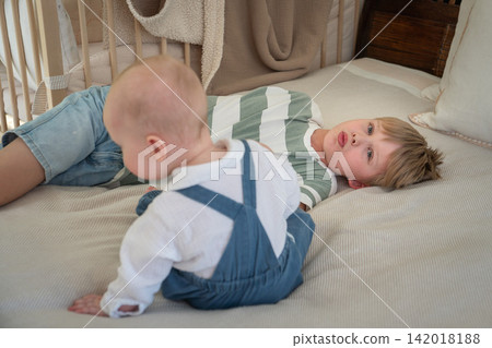 Baby sits on bed beside older brother while they relax and play together at home. Brotherhood, sibling bond, childhood, family connection, companionship, affection, play, development, togetherness 142018188