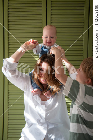 Mother carries baby son on her shoulders while older brother reaches up to play with him at home. Motherhood, sibling care, brotherhood, childhood, family bonding, play, affection, support Mother carries baby son on her shoulders while older brother reaches up to play with him at home. Motherhood, sibling care, brotherhood, childhood, family bonding, play, affection, support 142018189