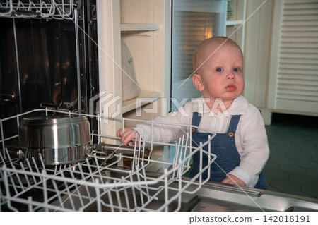 Baby stands beside open dishwasher and explores dish rack in home kitchen. Early childhood, curiosity, household safety, child development, exploration, learning, domestic life, everyday family Baby stands beside open dishwasher and explores dish rack in home kitchen. Early childhood, curiosity, household safety, child development, exploration, learning, domestic life, everyday family 142018191