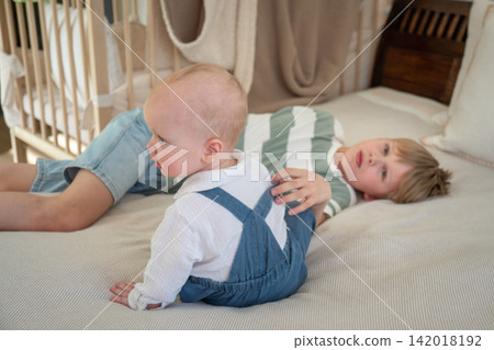 Baby sits on bed beside older brother while they relax and play together at home. Brotherhood, sibling bond, childhood, family connection, companionship, affection, play, development, togetherness 142018192