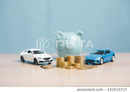 Piggy bank, white and blue toy cars, coins, and cash on desk, representing car savings, vehicle finance comparison, auto loan planning, purchase budget, and insurance concept. 142019727