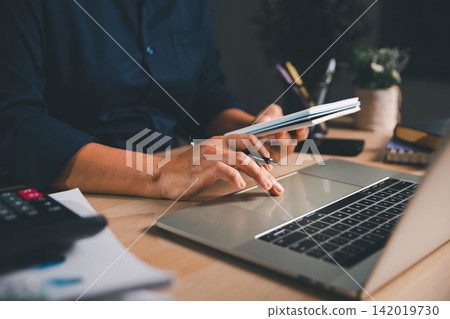 Business professional holding a notebook while using a laptop at a modern workspace, representing project planning, digital productivity, remote work, administration, and organized workflow. Business professional holding a notebook while using a laptop at a modern workspace, representing project planning, digital productivity, remote work, administration, and organized workflow. 142019730