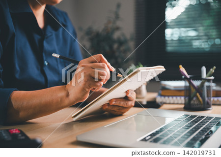 Professional holding a spiral notebook and pencil beside a laptop and calculator, representing business planning, budgeting, task management, financial organization, Professional holding a spiral notebook and pencil beside a laptop and calculator, representing business planning, budgeting, task management, financial organization, 142019731