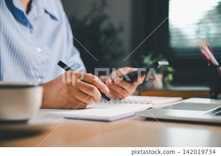 Person using a smartphone while writing notes beside a laptop, representing mobile learning, online research, multitasking, digital communication, remote work, and modern study planning. 142019734