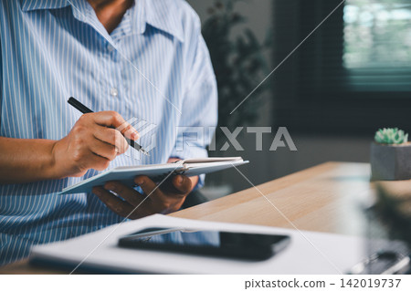 Person writing in a notebook while working beside a smartphone at a modern desk, representing daily planning, goal setting, note taking, personal organization, and productive remote work. 142019737