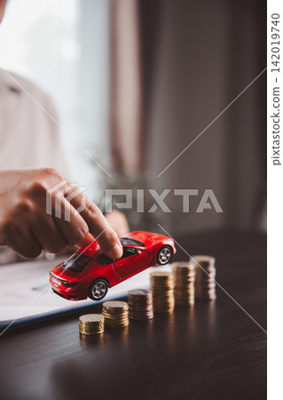 Hand lifts a red car model above rising coin stacks, representing progress toward vehicle ownership, auto savings, purchase goals, financing growth, investment planning, and financial achievement. Hand lifts a red car model above rising coin stacks, representing progress toward vehicle ownership, auto savings, purchase goals, financing growth, investment planning, and financial achievement. 142019740