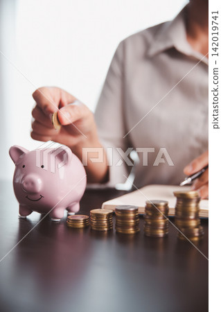 Person adding a coin to a pink piggy bank beside rising coin stacks and a notebook, illustrating regular saving, budget tracking, investment discipline, and steady measurable progress. Person adding a coin to a pink piggy bank beside rising coin stacks and a notebook, illustrating regular saving, budget tracking, investment discipline, and steady measurable progress. 142019741