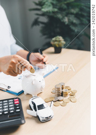 Person putting coin into piggy bank with calculator, white toy car, and coin stacks on desk, representing saving money for car purchase and auto loan planning. Person putting coin into piggy bank with calculator, white toy car, and coin stacks on desk, representing saving money for car purchase and auto loan planning. 142019742
