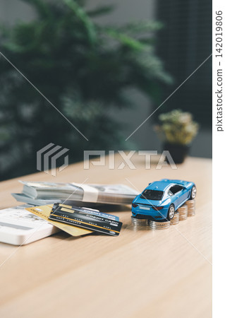 Blue miniature car on coin stacks with credit cards, cash, and calculator, showing vehicle financing, auto loan payment, car insurance, and personal budget planning. Blue miniature car on coin stacks with credit cards, cash, and calculator, showing vehicle financing, auto loan payment, car insurance, and personal budget planning. 142019806