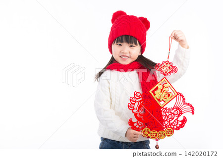 A happy little Asian girl in a red beanie holds a traditional Chinese New Year fish decoration against a white background. 142020172