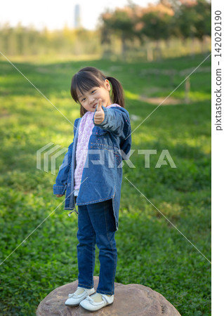 Joyful little Asian girl standing on a stone in the park giving thumbs up. Joyful little Asian girl standing on a stone in the park giving thumbs up. 142020190