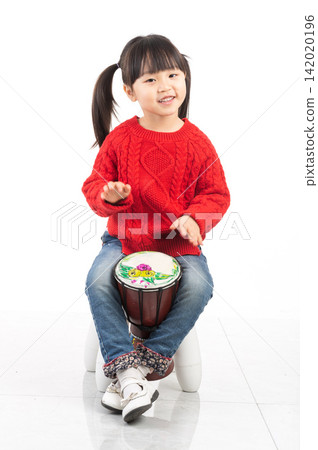 Smiling East Asian girl waving hand while playing an African djembe drum. Smiling East Asian girl waving hand while playing an African djembe drum. 142020196