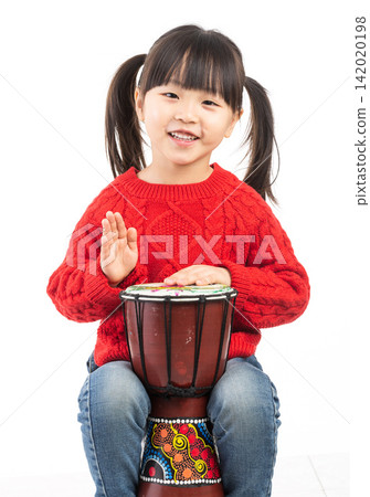 Smiling East Asian girl waving hand while playing an African djembe drum. Smiling East Asian girl waving hand while playing an African djembe drum. 142020198