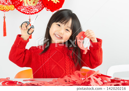A little Asian girl wearing a bright red sweater is intently cutting red paper, preparing to welcome the Lunar New Year. A little Asian girl wearing a bright red sweater is intently cutting red paper, preparing to welcome the Lunar New Year. 142020216