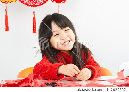 A little Asian girl wearing a bright red sweater is intently cutting red paper, preparing to welcome the Lunar New Year. 142020232
