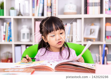 Attentive little student holding a pen and turning the page of a colorful textbook. 142020286