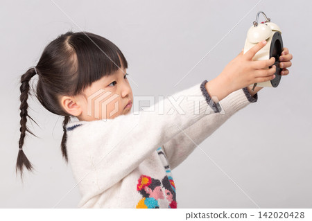 Side view of a little girl pushing a white alarm clock forward with both hands. Side view of a little girl pushing a white alarm clock forward with both hands. 142020428