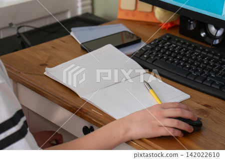 Schoolboy does homework at computer with open notebook, pencil and smartphone on wooden desk at home. Education, online learning, schoolwork, studying, concentration, technology, independent study Schoolboy does homework at computer with open notebook, pencil and smartphone on wooden desk at home. Education, online learning, schoolwork, studying, concentration, technology, independent study 142022610