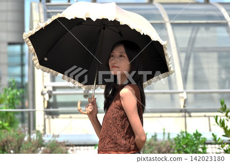 一位年輕女子麵帶微笑,在有花壇的戶外露臺上撐著陽傘(夏季,防曬)。 一位年輕女子麵帶微笑,在有花壇的戶外露臺上撐著陽傘(夏季,防曬)。 142023180