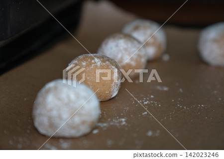 Raw almond cookie dough balls coated with powdered sugar rest on baking paper before baking. Home baking, dessert preparation, confectionery, handmade pastry, recipe, baking process Raw almond cookie dough balls coated with powdered sugar rest on baking paper before baking. Home baking, dessert preparation, confectionery, handmade pastry, recipe, baking process 142023246