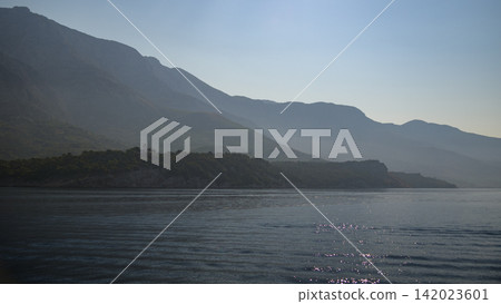 A serene coastal landscape viewed from a body of water, featuring a deep blue sea reflecting sunlight on ripples and peaks of gray mountains in a hazy misty atmosphere A serene coastal landscape viewed from a body of water, featuring a deep blue sea reflecting sunlight on ripples and peaks of gray mountains in a hazy misty atmosphere 142023601