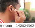 Man gently cleans ear with cotton swab. Focused expression suggests care and hygiene. Outdoor setting with soft natural light. Ideal for health, wellness, or personal care content 142024302