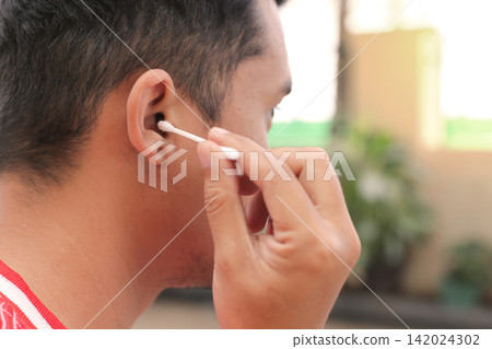 Man gently cleans ear with cotton swab. Focused expression suggests care and hygiene. Outdoor setting with soft natural light. Ideal for health, wellness, or personal care content Man gently cleans ear with cotton swab. Focused expression suggests care and hygiene. Outdoor setting with soft natural light. Ideal for health, wellness, or personal care content 142024302